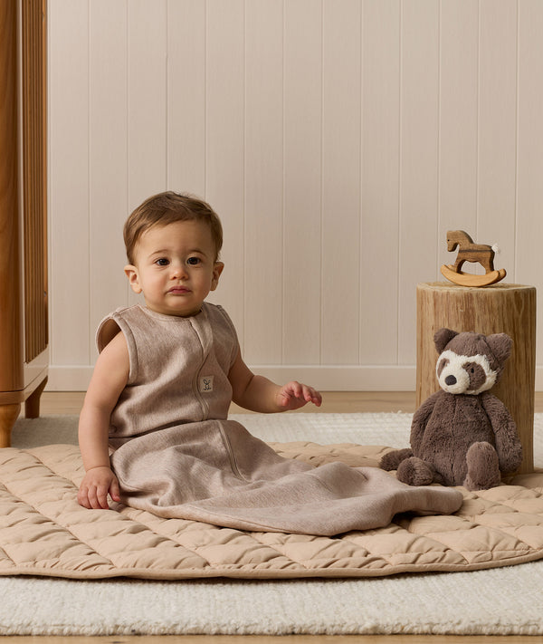 Infant wearing a sleeveless Sleeping Bag 1.0 TOG, sitting next to a plush toy on a quilted mat.