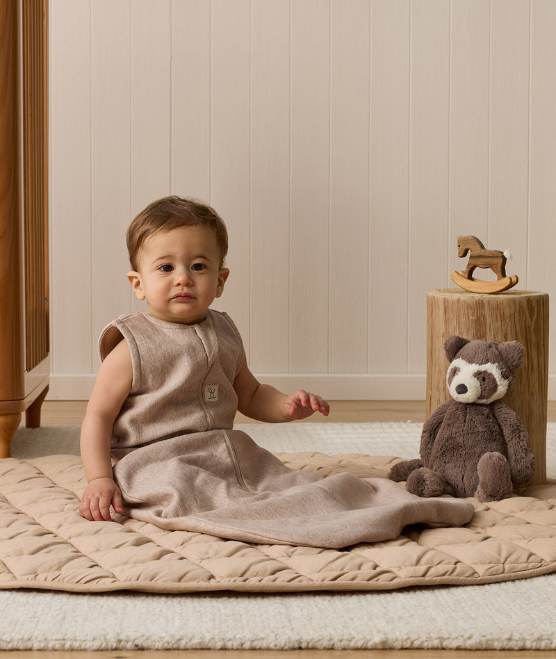 Infant wearing a sleeveless Sleeping Bag 1.0 TOG, sitting next to a plush toy on a quilted mat.