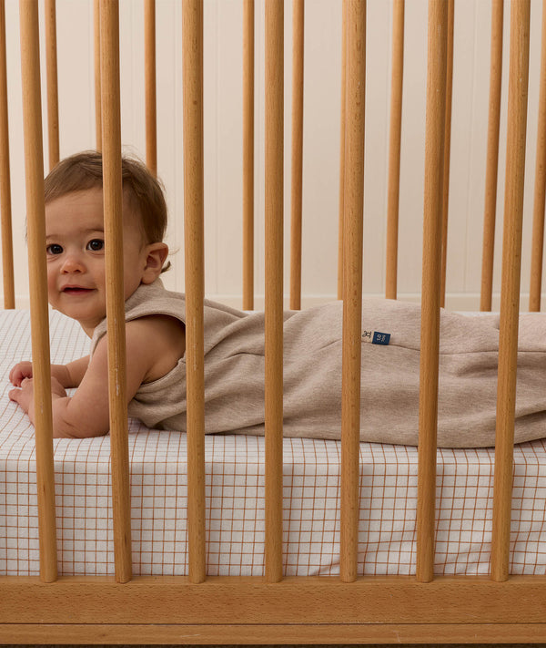 A smiling baby wearing a Taupe Marle Sleeping Bag, lying on their stomach inside a wooden cot with a grid-patterned sheet.