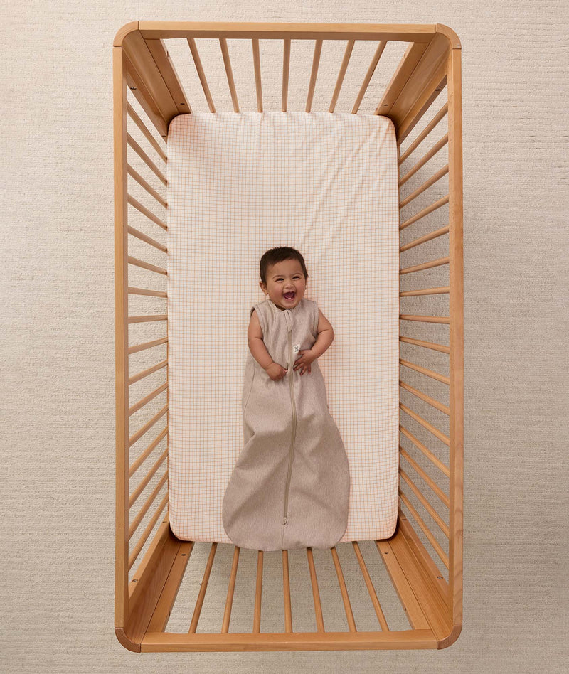 A happy baby lying in a wooden cot wearing a sleeveless Sleeping Bag 1.0 TOG Taupe Marle
