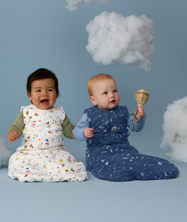 Two babies wearing ergoPouch Sleeping Bags in Daydream and Starbeam prints, sitting side by side against a blue background with decorative clouds above.