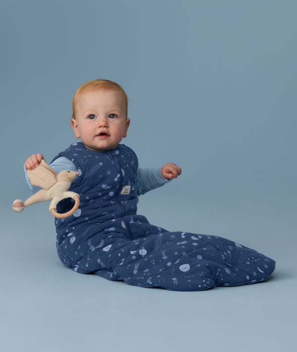 Baby wearing a blue ergoPouch Sleeping Bag 2.5 TOG, sitting against a blue background while holding a toy