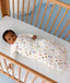 Baby lying in a wooden cot wearing a white sleeved Sleeping Bag with colourful print