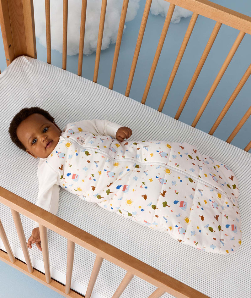 Baby lying in a wooden cot wearing a white sleeved Sleeping Bag with colourful print