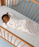 Baby lying in a wooden cot wearing a white Sleeping Bag 3.5 TOG, featuring colourful all-over print and long sleeves.