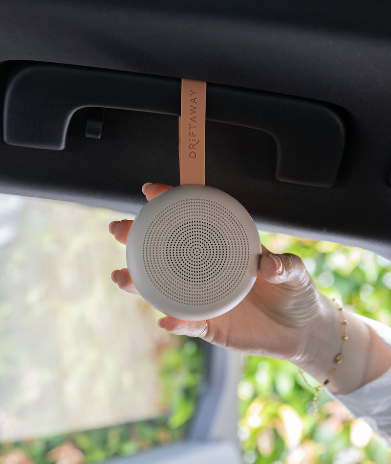A hand holds a small round white noise machine hanging from a tan strap labeled, attached to a car visor.