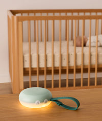 A soft green white noise machine with a glowing warm night light sits on a wooden table in the foreground. It features buttons, including music and power icons, and a green strap with the brand name “Drift Away.” In the background, a baby is peacefully sleeping in a wooden crib, emphasizing the calming effect of the device.