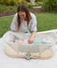 Portable bassinet set up outdoors on grass, featuring a foldable frame and mesh side panels with a baby lying inside and a mother reaching in.