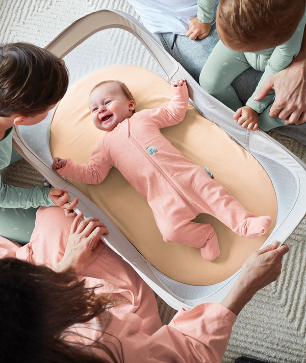 A smiling baby wearing a pink long sleeve layer lies happily inside a portable mesh-sided bassinet surrounded by family.