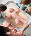 A smiling baby wearing a pink long sleeve layer lies happily inside a portable mesh-sided bassinet surrounded by family.