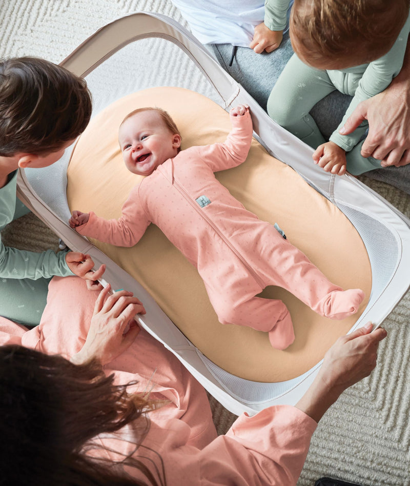 A smiling baby wearing a pink long sleeve layer lies happily inside a portable mesh-sided bassinet surrounded by family.