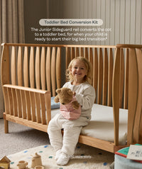 Child sitting in a wooden cot with a teddy bear, promoting a toddler bed conversion kit.
