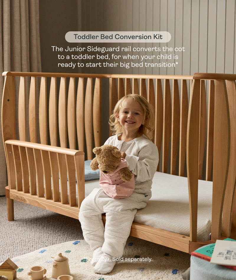 Child sitting in a wooden cot with a teddy bear, promoting a toddler bed conversion kit.