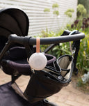 A compact white noise machine by from ergoPouch with a circular speaker design is attached to a stroller handle using a tan leather strap embossed with the name “Drift Away.”