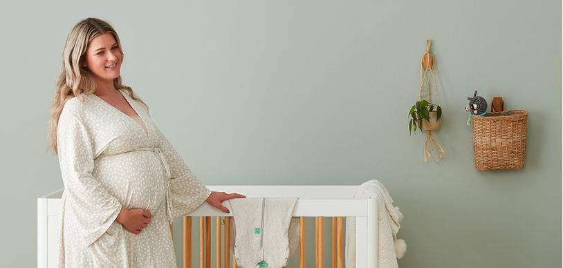 Pregnant woman standing next to crib, planning what to pack in her baby hospital bag, wearing ergoPouch Matchy Matchy Robe