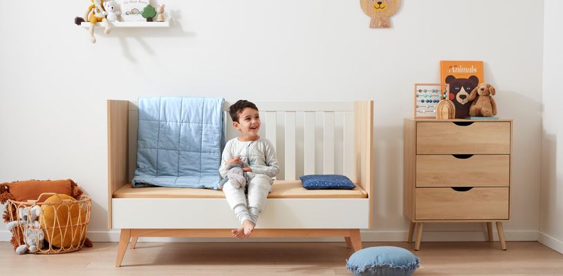 Boy sitting on his cot with one side down to begin mimicking the experience of a big bed before the transition from cot to bed