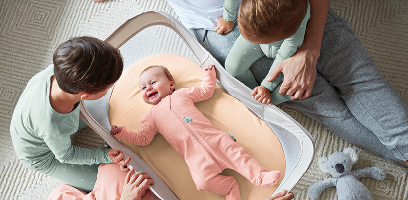 Two young children lean over the Easy Sleep Portable Baby Bassinet inside of which baby is smiling