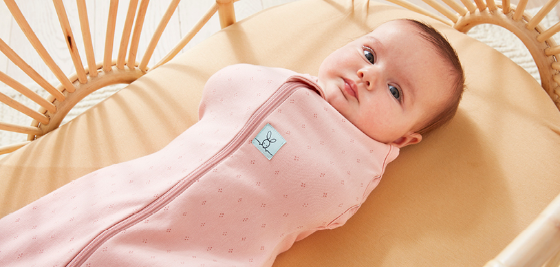 Baby swaddled in pink ergoPouch swaddle bag, resting in bassinet