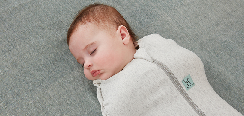 Newborn baby sleeping restfully on back, wearing a swaddle pouch