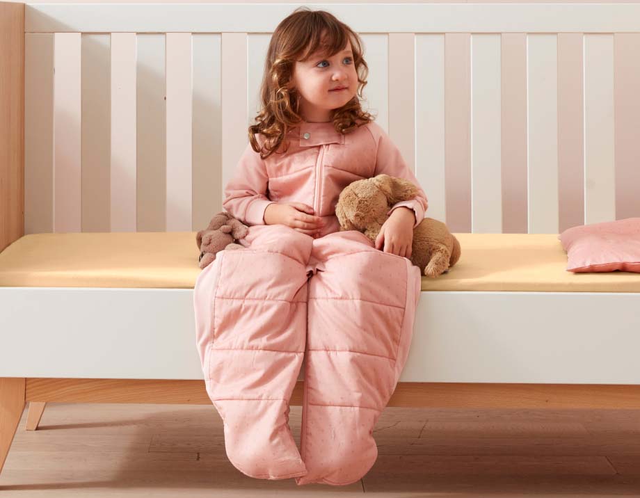 Toddler sitting on edge of bed wearing Sleep Suit Bag in Berries, holding a teddy